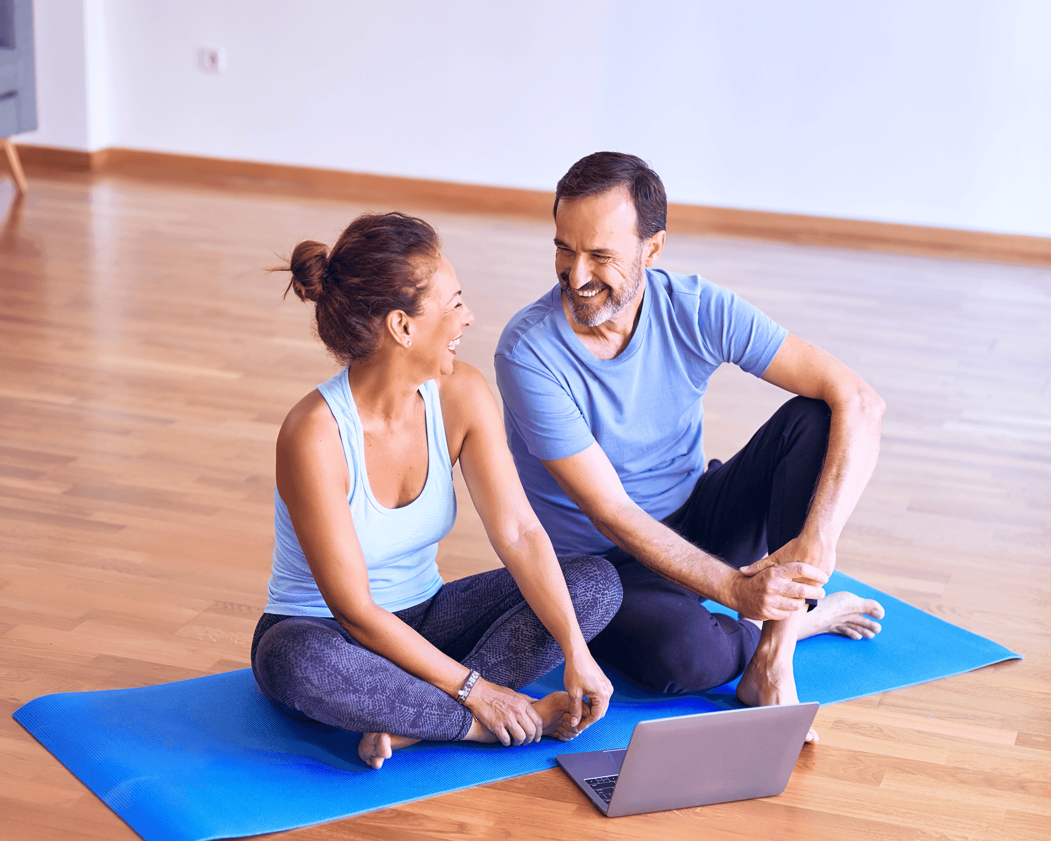 Eine Frau und ein Mann sitzen auf einer blauen Yogamatte in einem Turnraum und schauen sich auf einem Laptop Rehaübungen an.